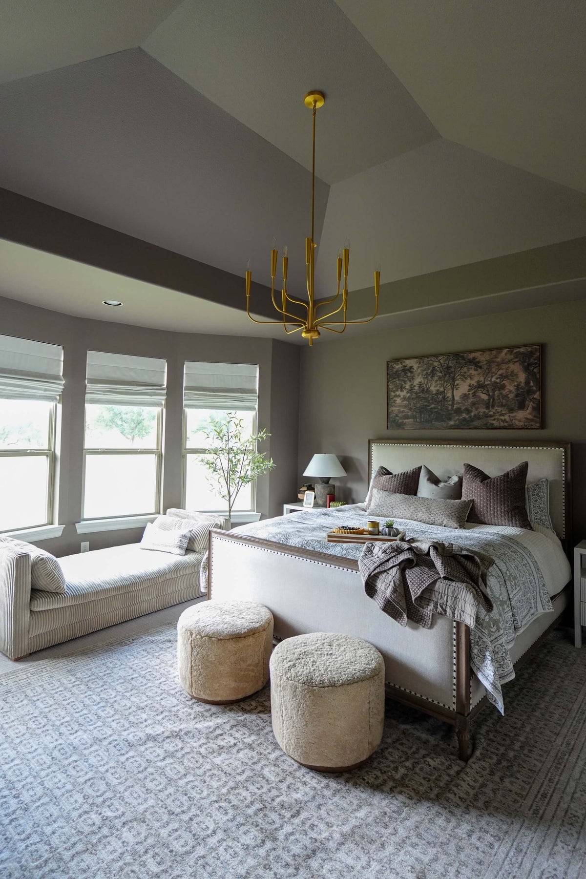 A styled bedroom interior with a bed dressed in neutral bedding, two round pouf ottomans at the foot of the bed, a chaise lounge by large windows, and a gold chandelier overhead.