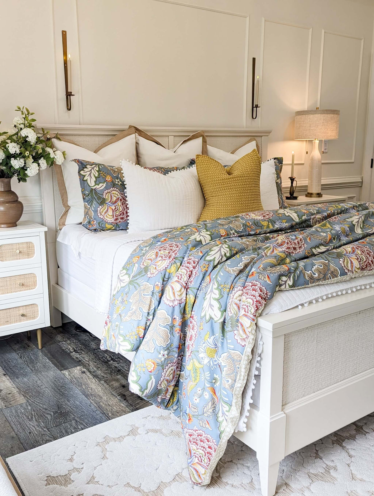 A styled bedroom scene featuring a white wooden bed dressed with multiple decorative pillows and a light blue floral patterned quilt. The room has cream paneled walls, a white nightstand with flowers, a lit table lamp, and dark wood flooring.