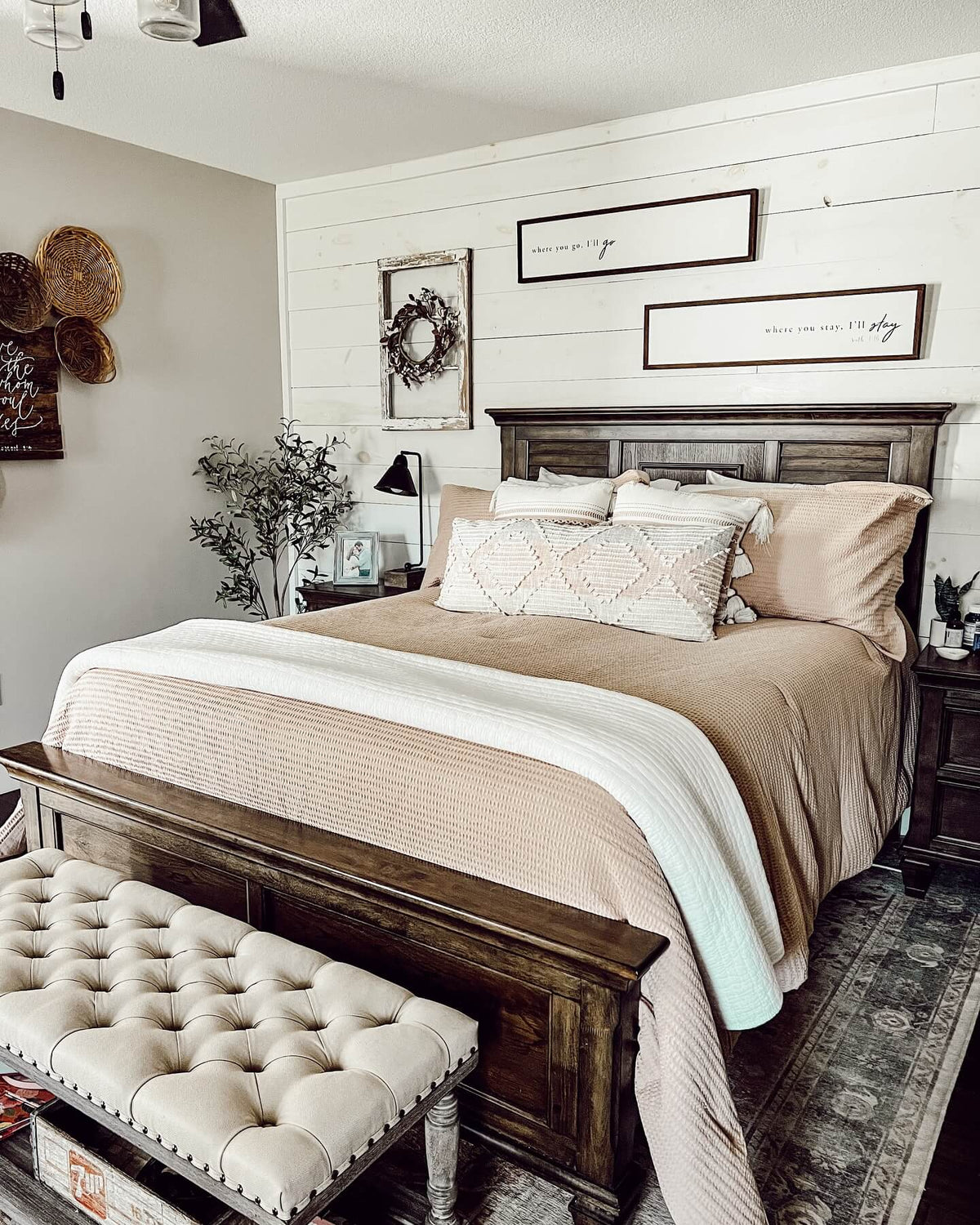 A styled bedroom scene featuring a bed dressed in beige and cream bedding, a dark wood headboard and footboard, a tufted cream bench at the foot of the bed, and a shiplap wall with framed wall art behind it.