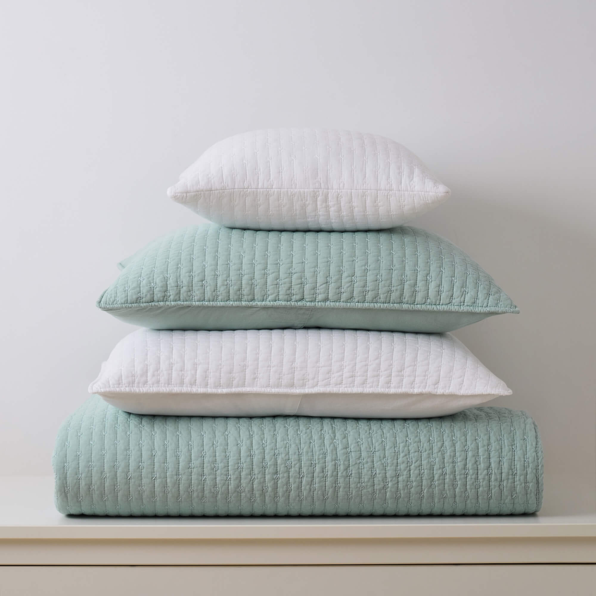 Stack of light blue quilt, white pillow, and light blue pillow on a white surface with a light gray background