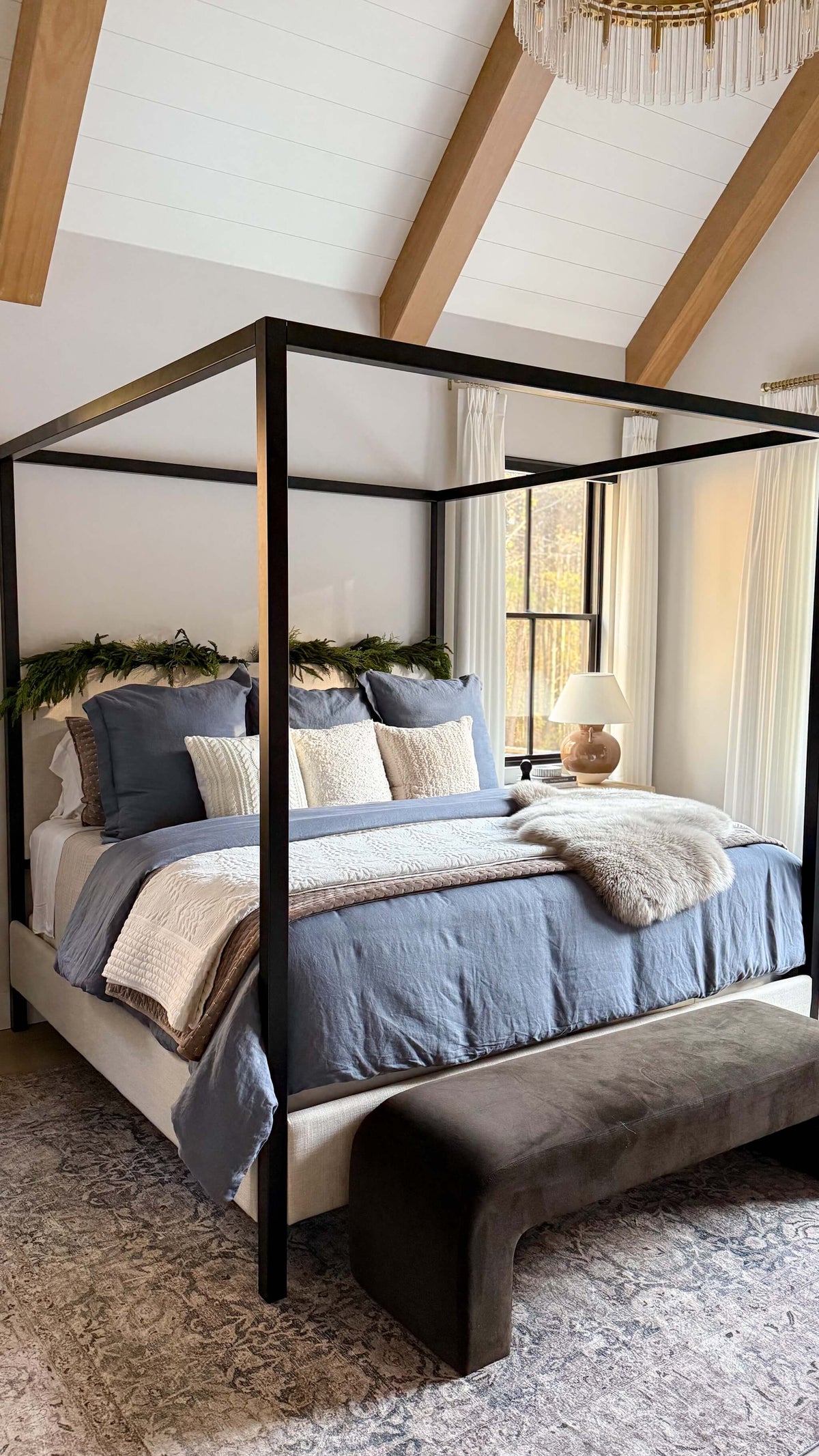 A rustic-style four-poster canopy bed with a black metal frame, dressed in slate blue and beige bedding, set in a bedroom with a wooden bench at the foot of the bed.