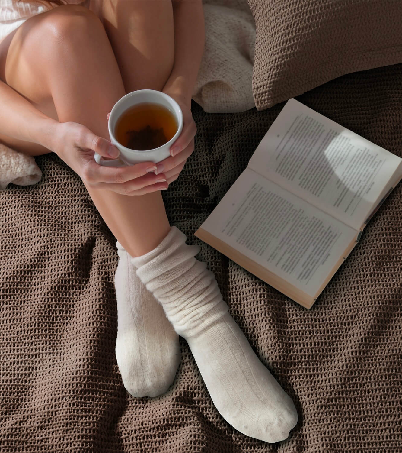 Person sitting on a textured duvet holding a cup of tea with an open book beside them.