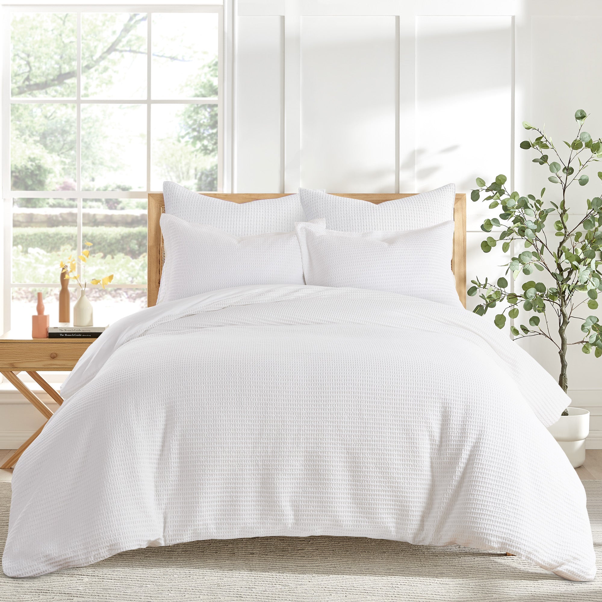 A neatly made bed with a white textured cotton comforter set and matching shams, styled in a bright bedroom with a wooden headboard, a potted plant, and a side table by a large window