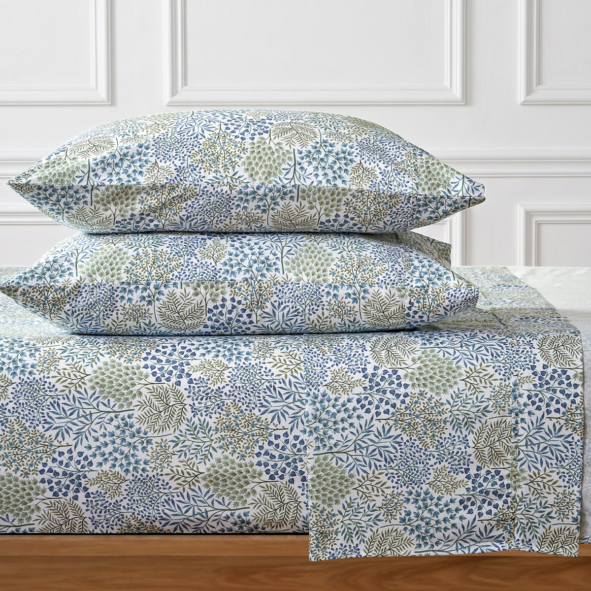 A bed dressed in a patterned sheet and two matching pillowcases, featuring a botanical floral print in blue, sage green, and beige on a white background, set against a white paneled wall.