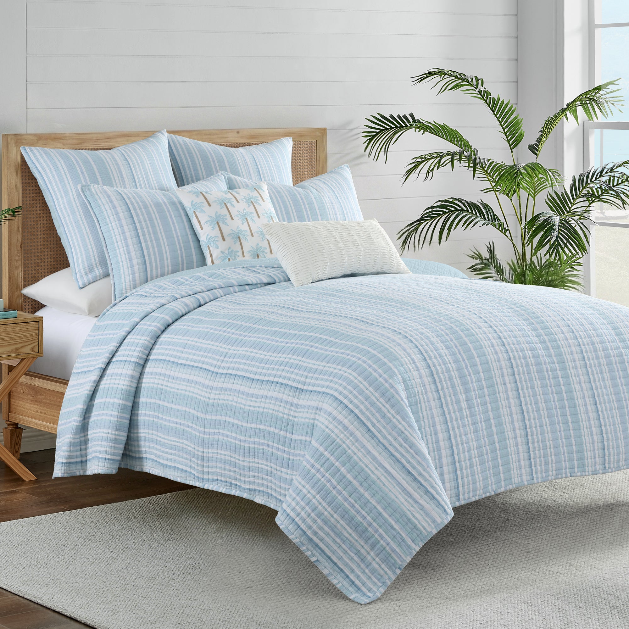 Boracay Stripe Crewel Palm Tree Pillow on a bed with light blue striped bedding and tropical decor.