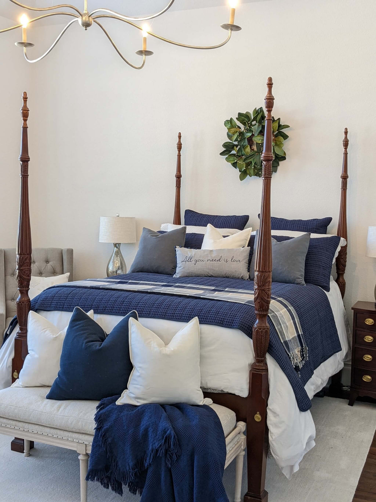 A styled bedroom scene featuring a dark wood four-poster bed dressed in navy blue and white bedding, with matching pillows, a bench at the foot of the bed, and a green wreath on the wall.