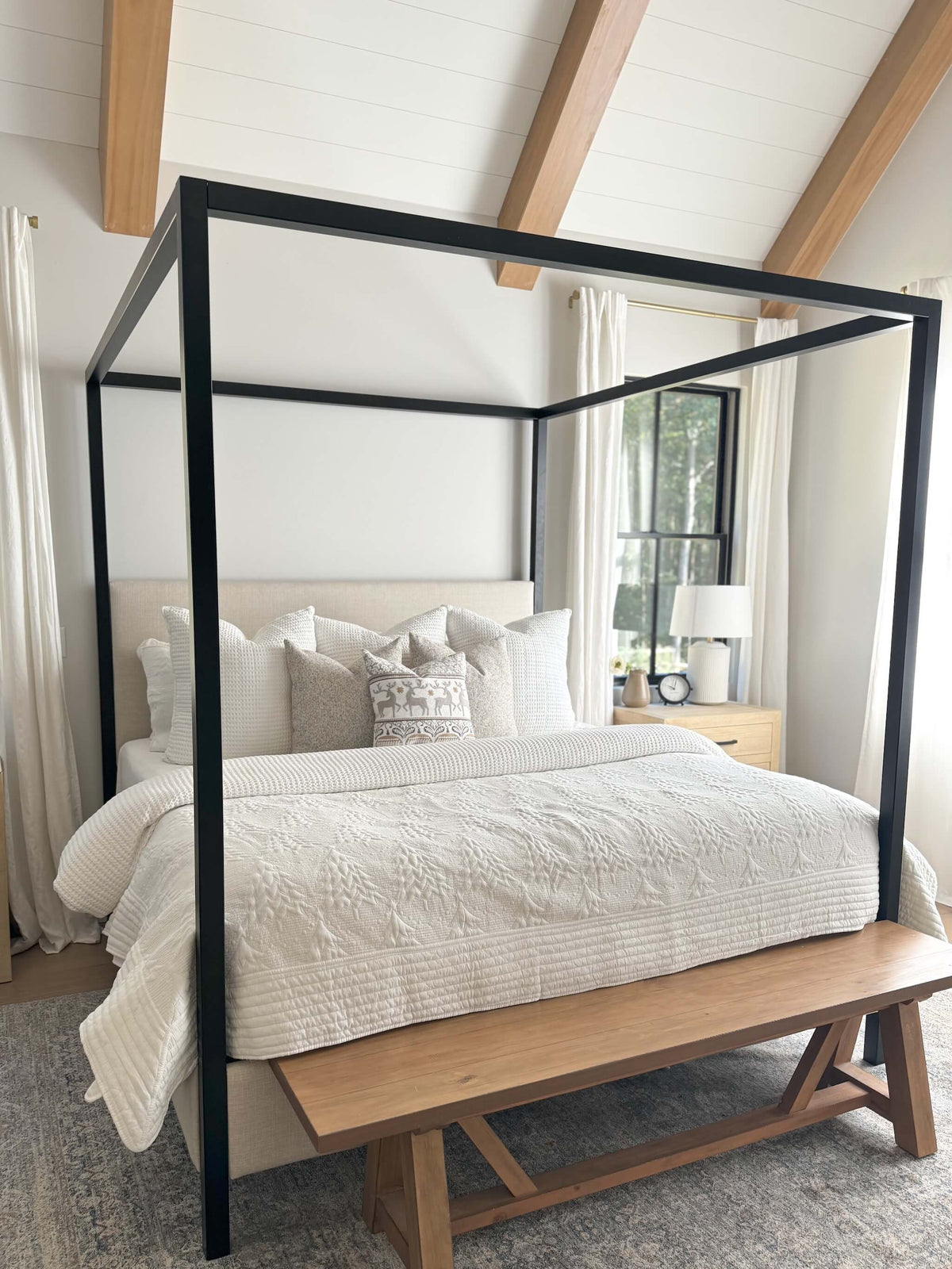 A four-poster canopy bed with a black metal frame and upholstered beige headboard, dressed in white textured bedding, set in a bright bedroom with a wooden bench at the foot of the bed.