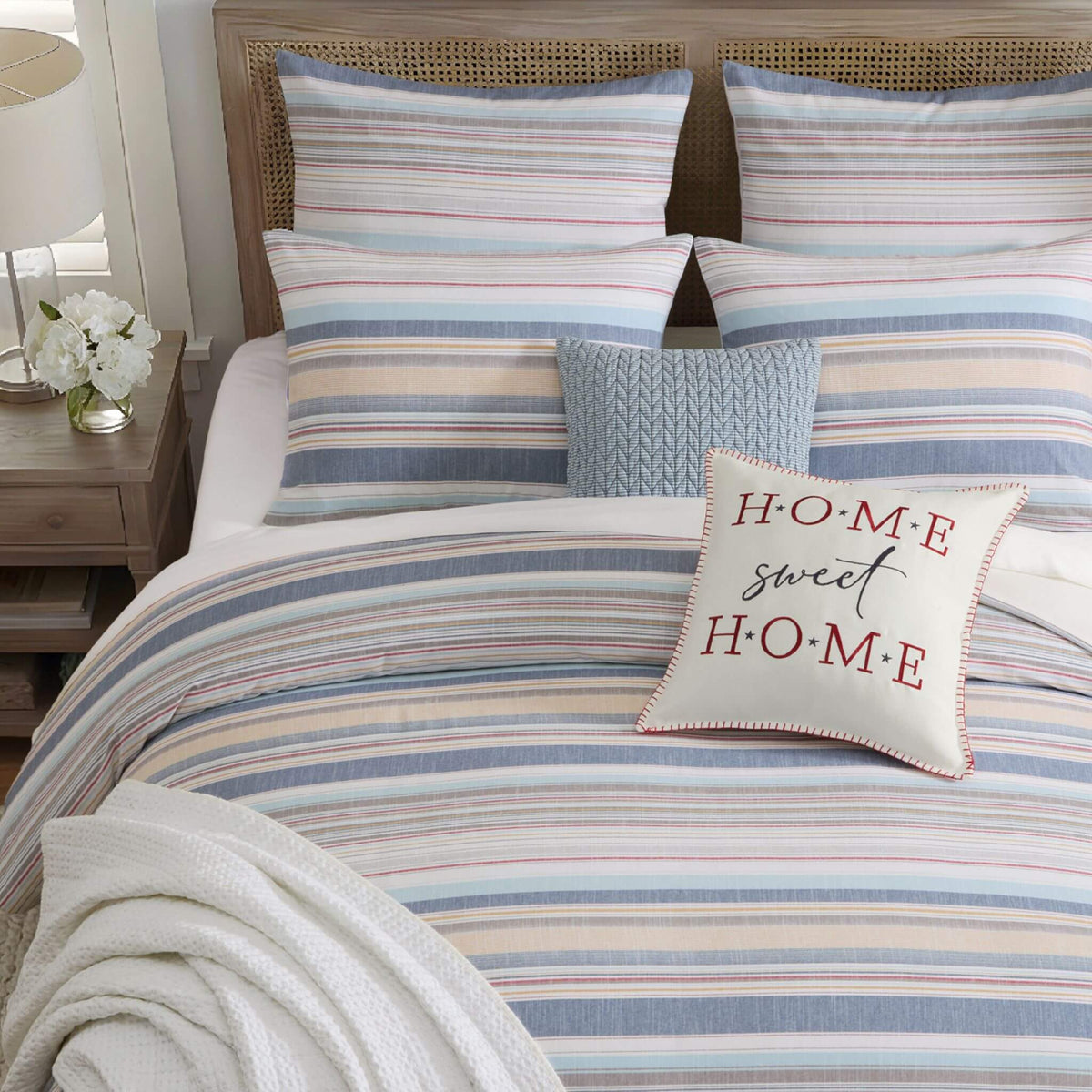 A neatly made bed with a striped bedding set in soft blue, beige, and white tones, including matching pillow shams and a textured light blue accent pillow. A cream throw pillow with the words 'HOME sweet HOME' in red script and block letters rests on the duvet.