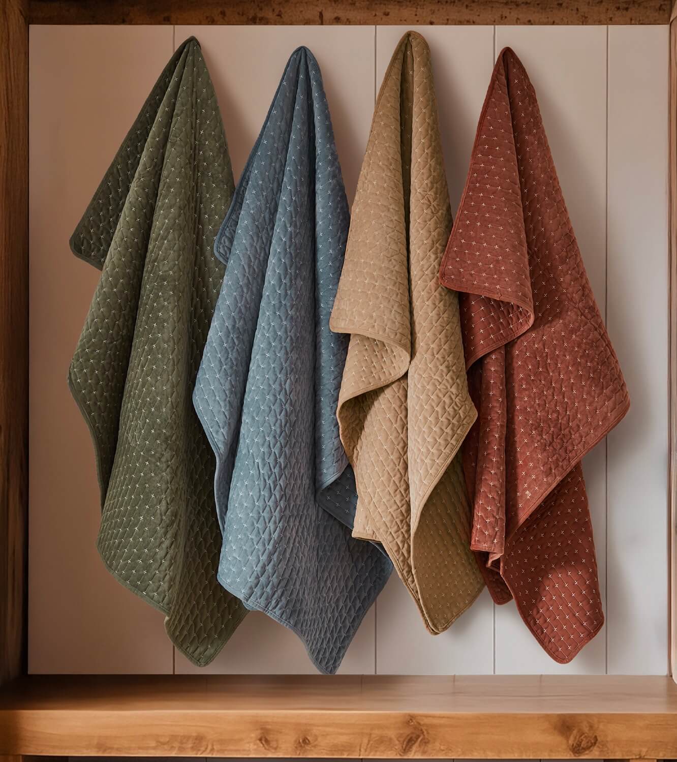 Four textured quilted throws in green, blue, beige, and rusty red hanging on a wooden shelf.