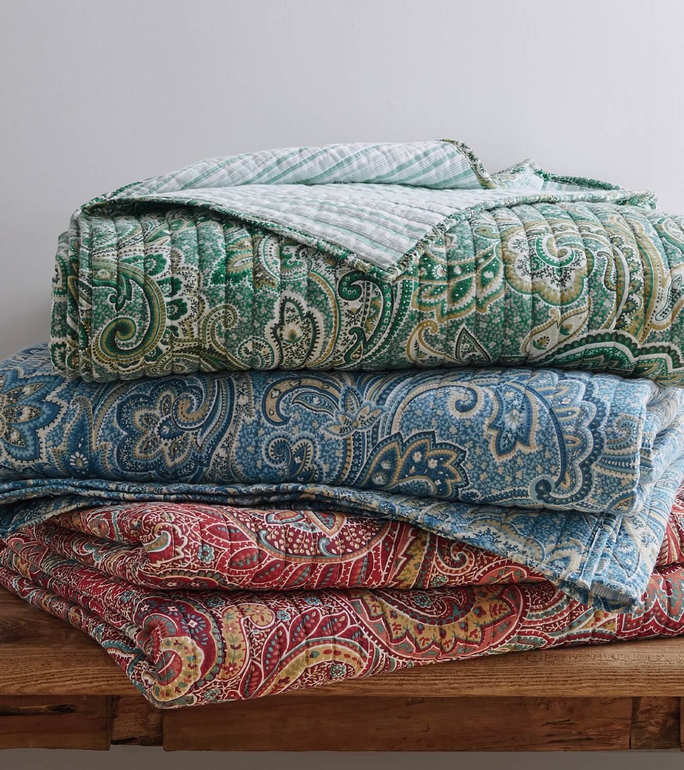 Stack of paisley-patterned quilts in green, blue, and red on a wooden surface.