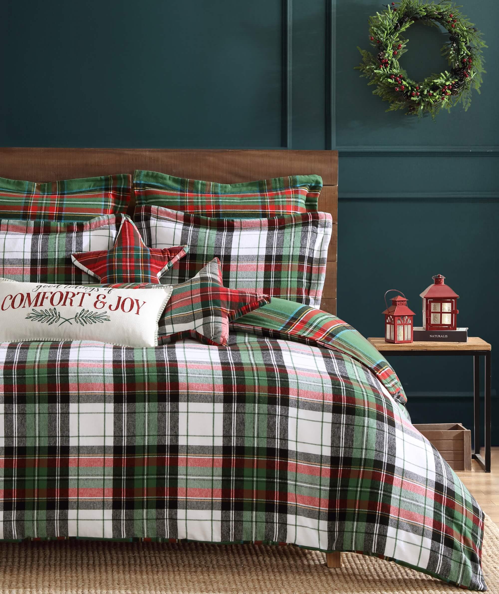 Plaid bedding set in red, green and white colors with festive pillows and a wreath on a dark green wall.
