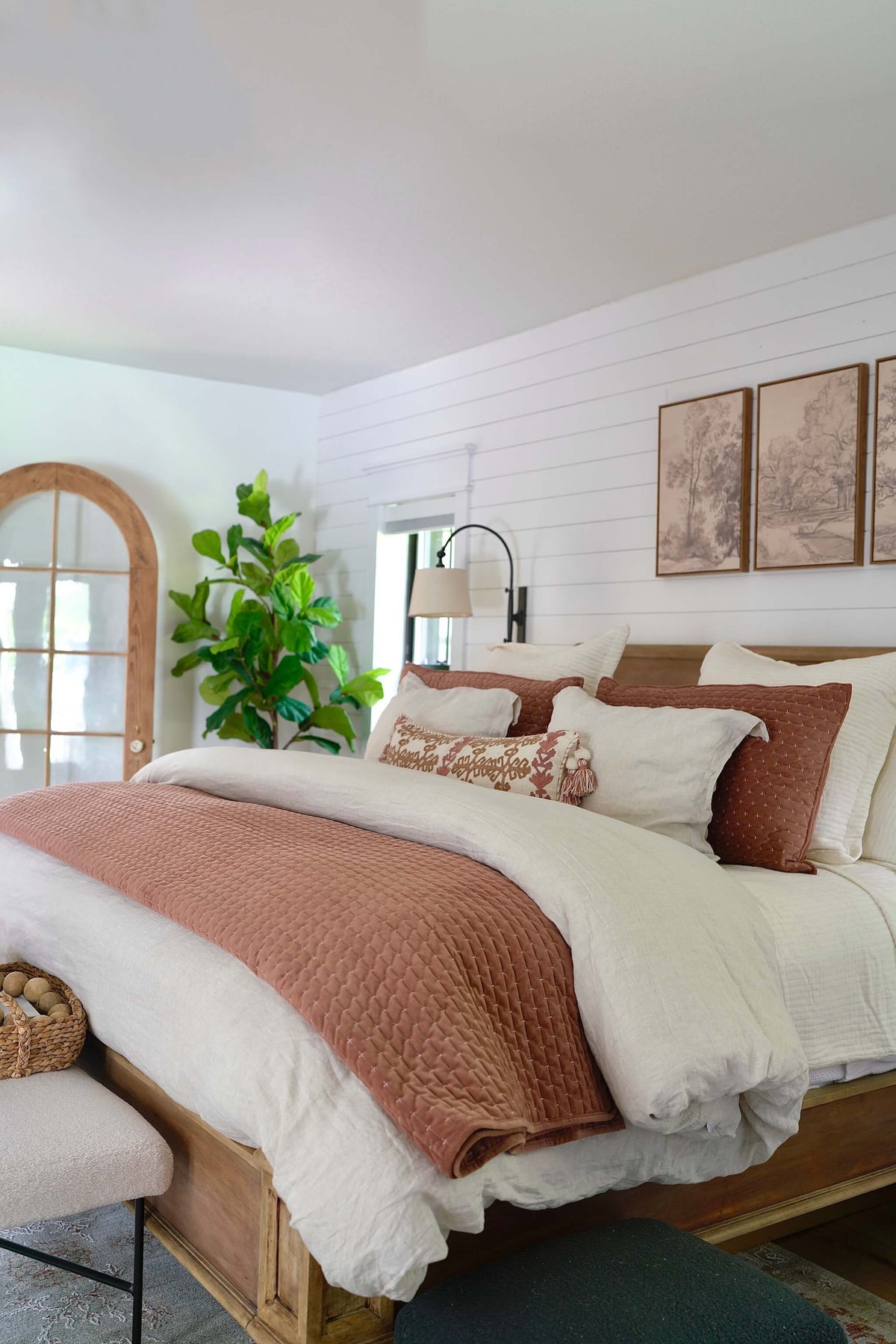 A styled bedroom scene featuring a wooden bed frame dressed in white bedding with a rust-brown quilted throw blanket and matching pillows. The background has white shiplap walls, framed art, a green plant, and a window with a wooden arch frame.