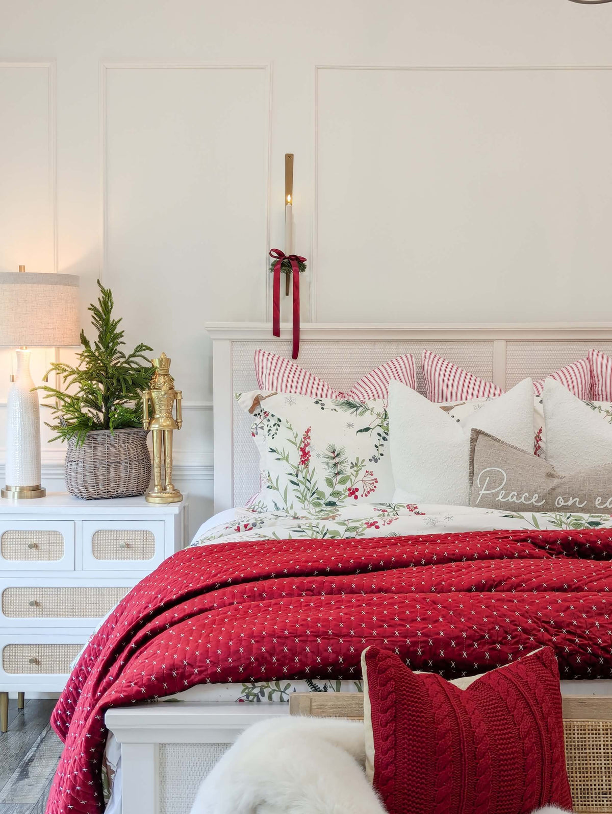 A styled bedroom scene featuring a bed dressed in a red patterned blanket, white and red striped pillows, and botanical patterned accent pillows. The bed is flanked by white nightstands with a lamp, a potted plant, and decorative figurines, set against a white paneled wall.