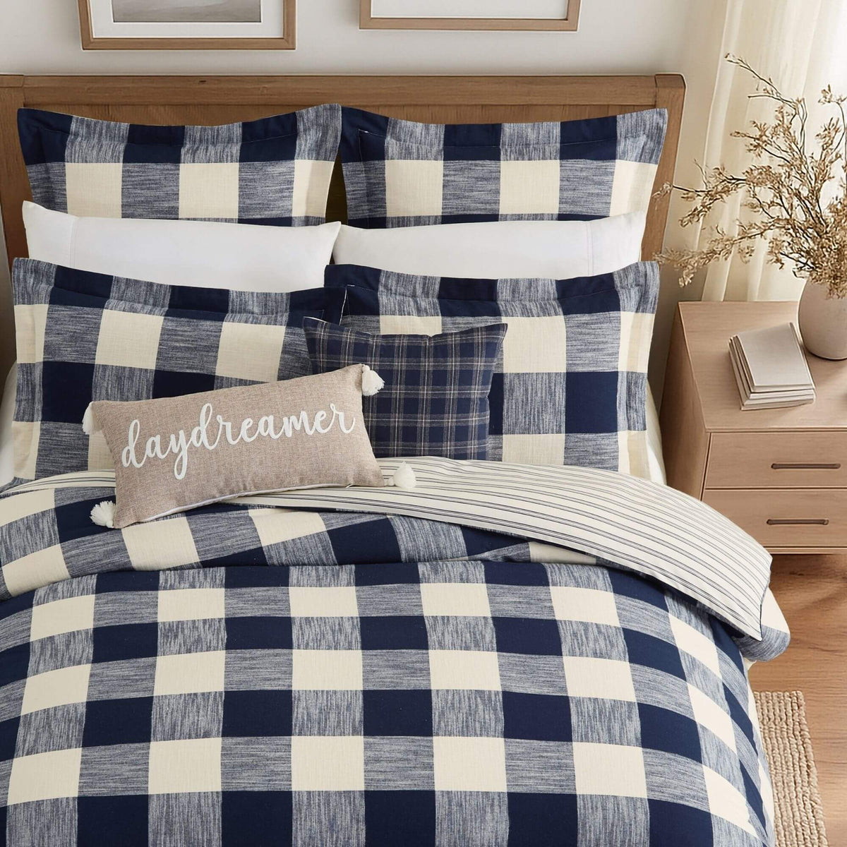 A bed styled with a navy and cream buffalo plaid bedding set, including a comforter, matching pillow shams, a striped accent pillow, and a beige throw pillow with the word 'daydreamer' in white script.
