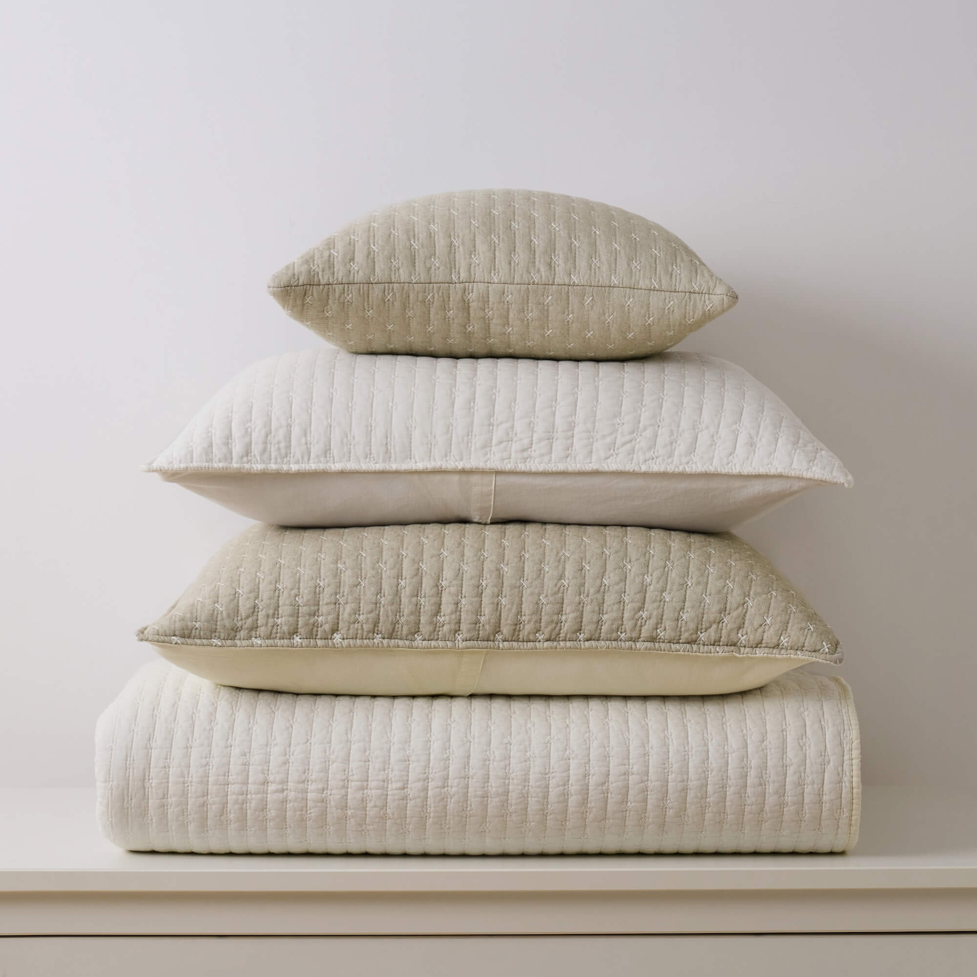 Stack of beige textured pillows and quilt on a white surface with a light gray background