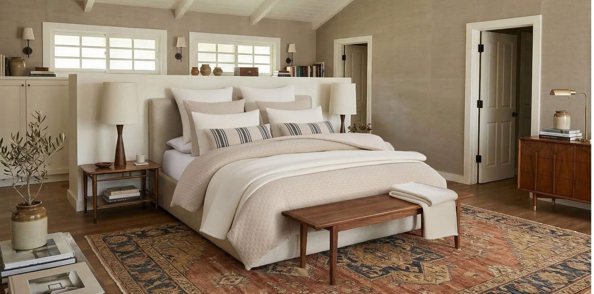 A styled bedroom interior featuring a bed dressed in neutral beige and cream bedding, with striped accent pillows, a wooden bench at the foot of the bed, matching nightstands, lamps, a patterned rug, and warm neutral walls.