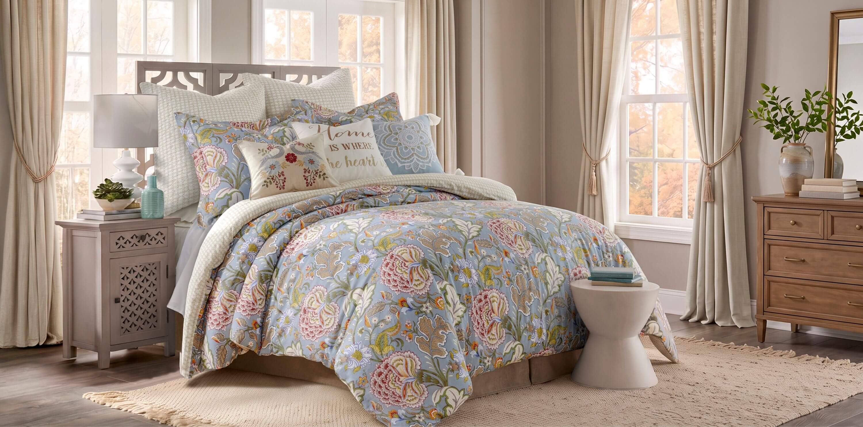 A floral comforter set styled on a bed in a bright, neutral-toned bedroom. The comforter features a light blue base with pink, green, and yellow floral and paisley patterns, accompanied by matching patterned pillows and shams.