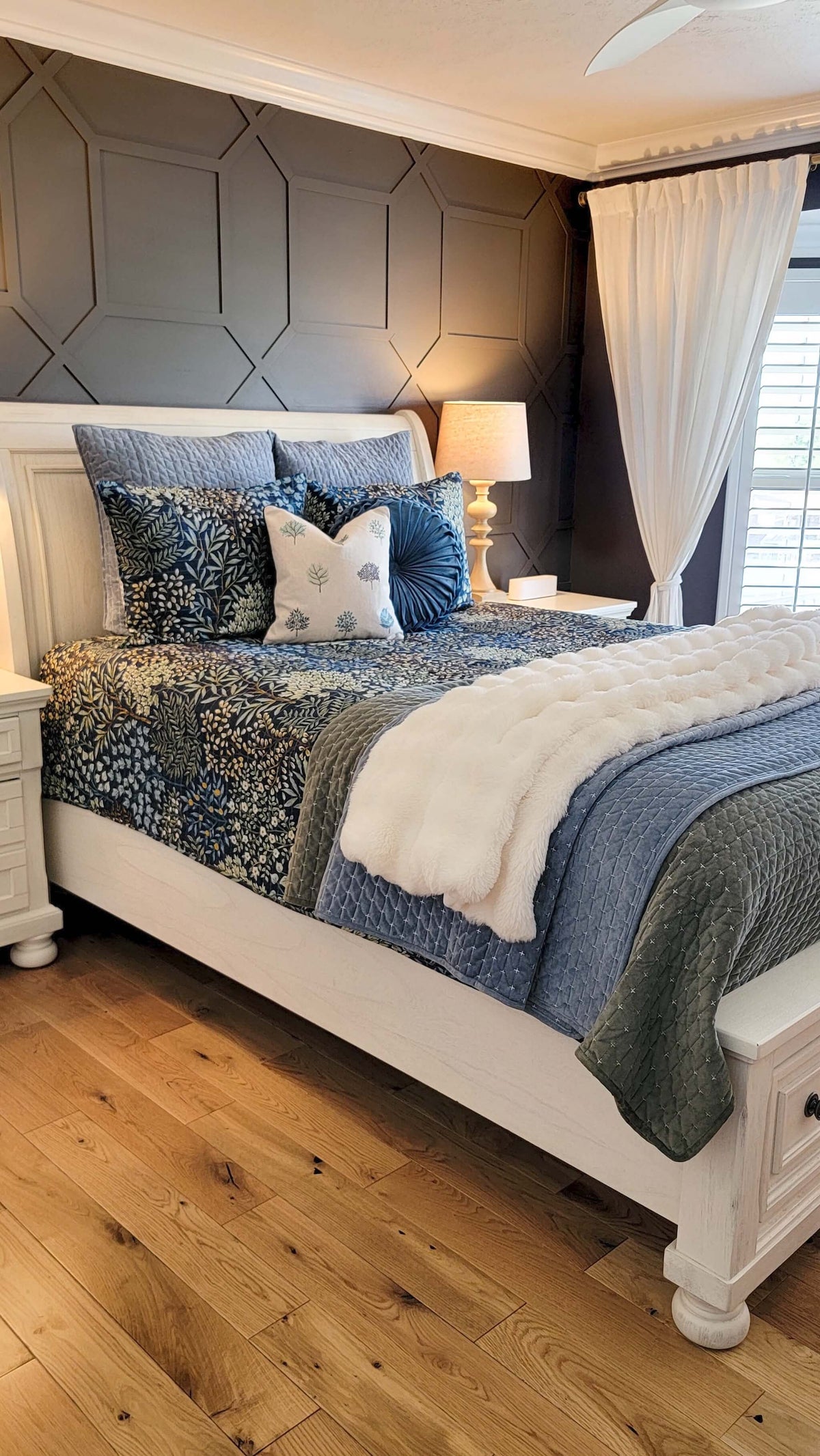 A styled bedroom scene featuring a white wooden bed frame dressed in blue and gray patterned bedding, including a textured blue quilt, patterned pillows, and a white throw blanket. The background includes a gray geometric paneled wall, white curtains, and light wood flooring.