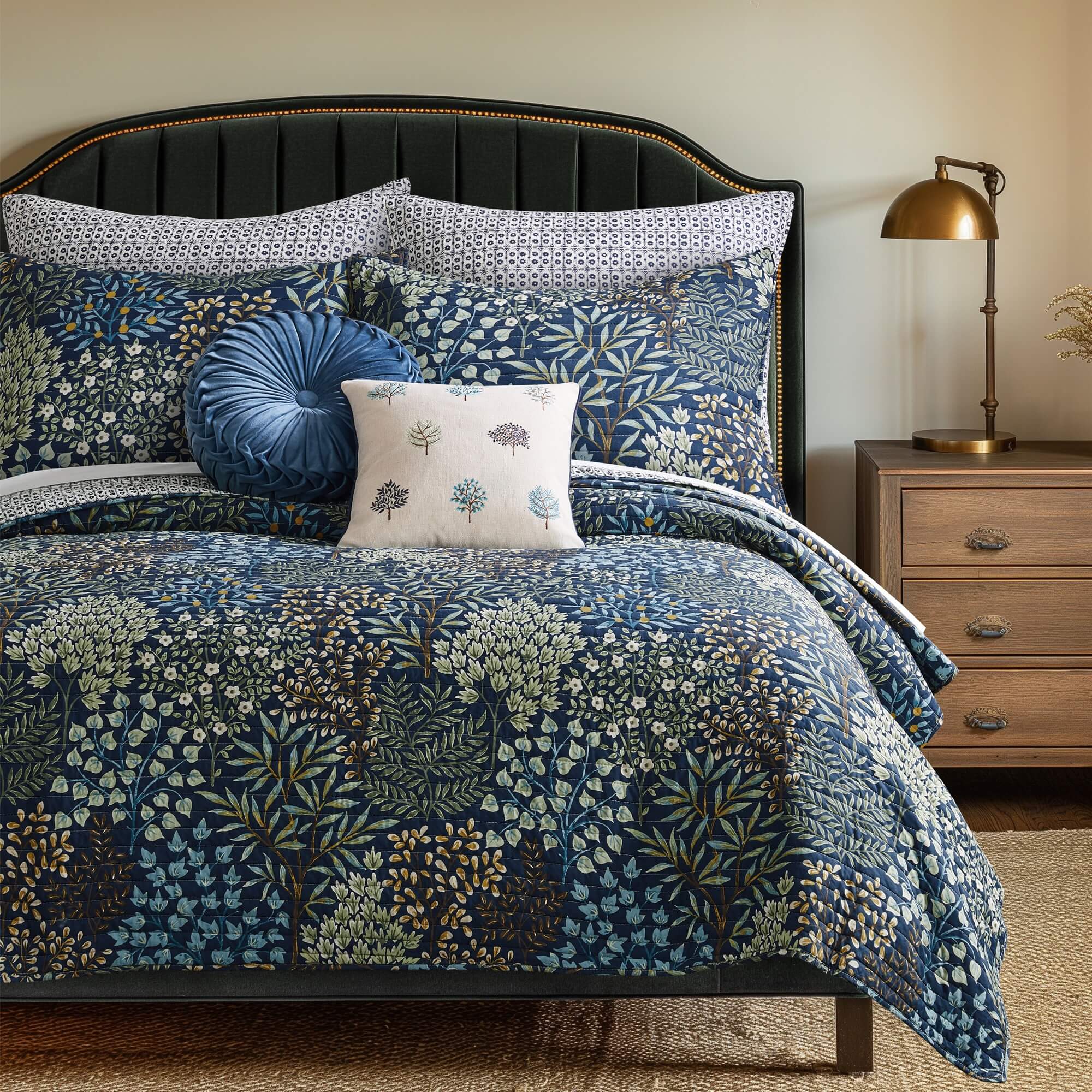 Blue floral bedding set with patterned shams, decorative pillows, and a wooden nightstand with brass lamp in a cozy bedroom.