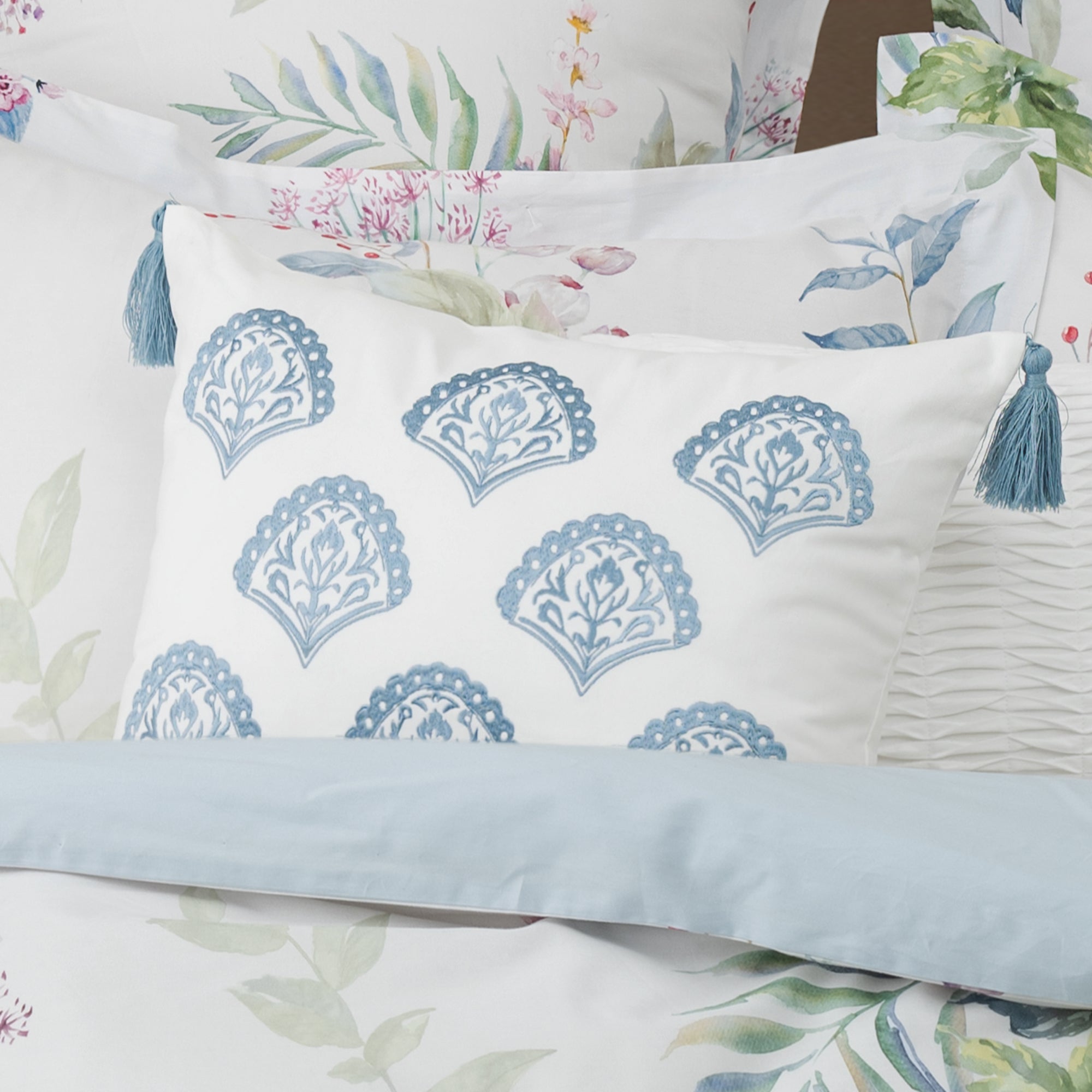 Cabana Stripe Blue Embroidered Medallion Pillow with blue tassels on a bed with floral bedding.