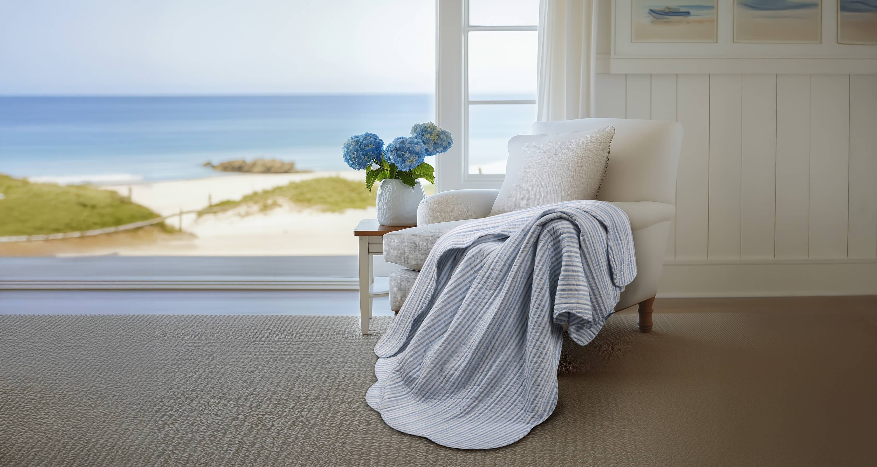 Cozy living room with a white armchair, blanket, and vase of flowers, overlooking a beach.