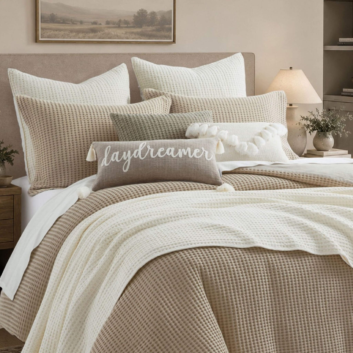 A styled bed in a warm neutral bedroom setting, featuring a beige and cream waffle-weave bedding set with textured pillows, a cream knit throw blanket, and a decorative pillow embroidered with the word 'dreamer'.