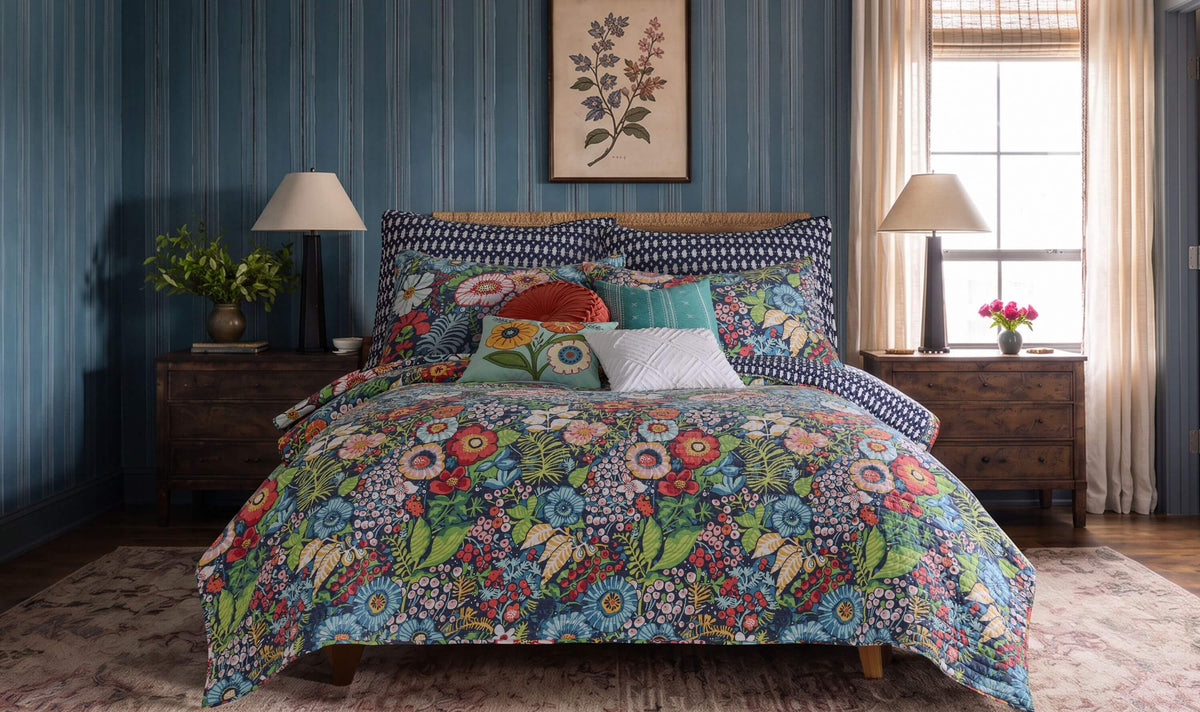 A bed dressed in a colorful floral and botanical print comforter set with matching pillows, styled in a bedroom with blue striped wallpaper, wooden nightstands, lamps, and a window.