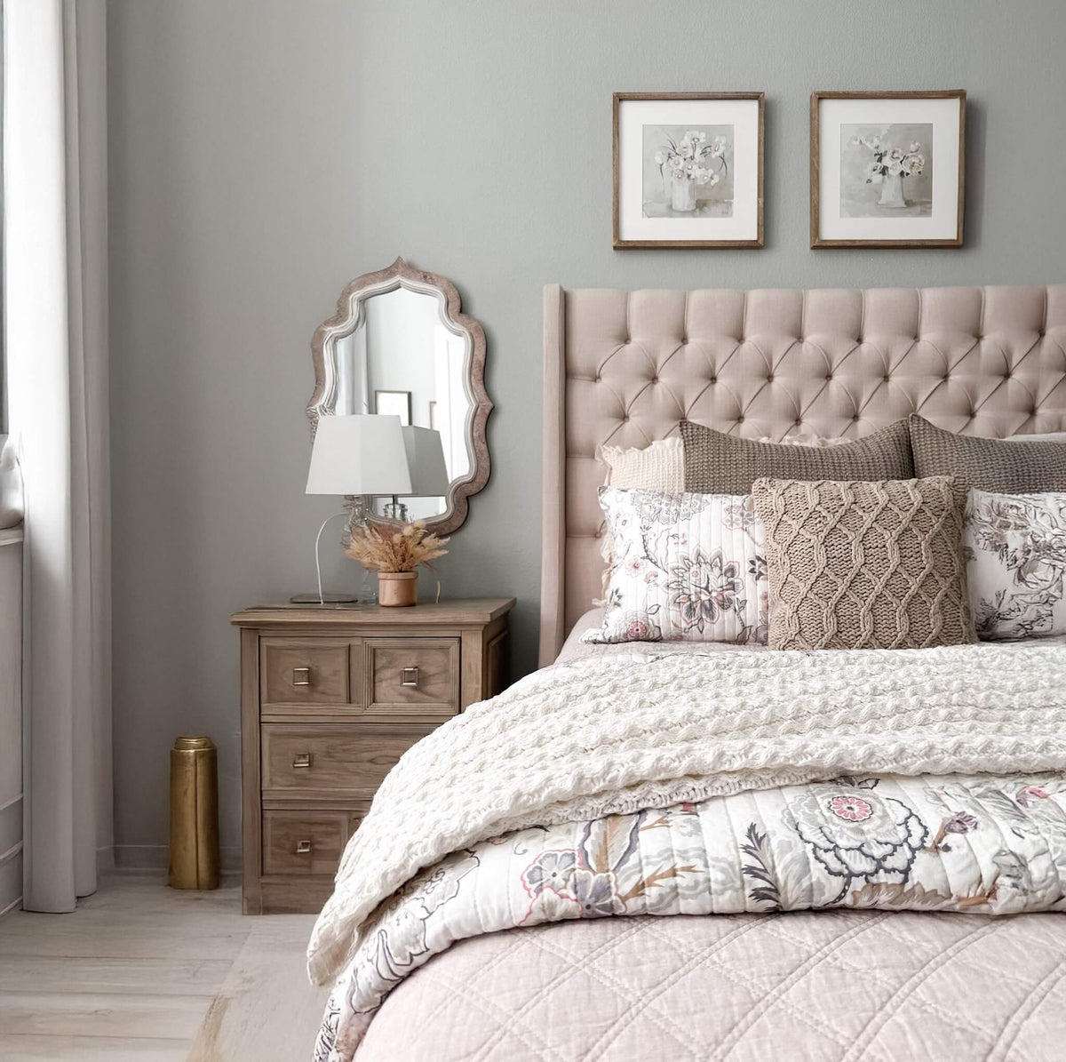 A styled bedroom scene featuring a bed with a tufted blush pink headboard, patterned bedding, matching pillows, a wooden nightstand, an ornate wall mirror, and two framed floral prints on a light gray wall.