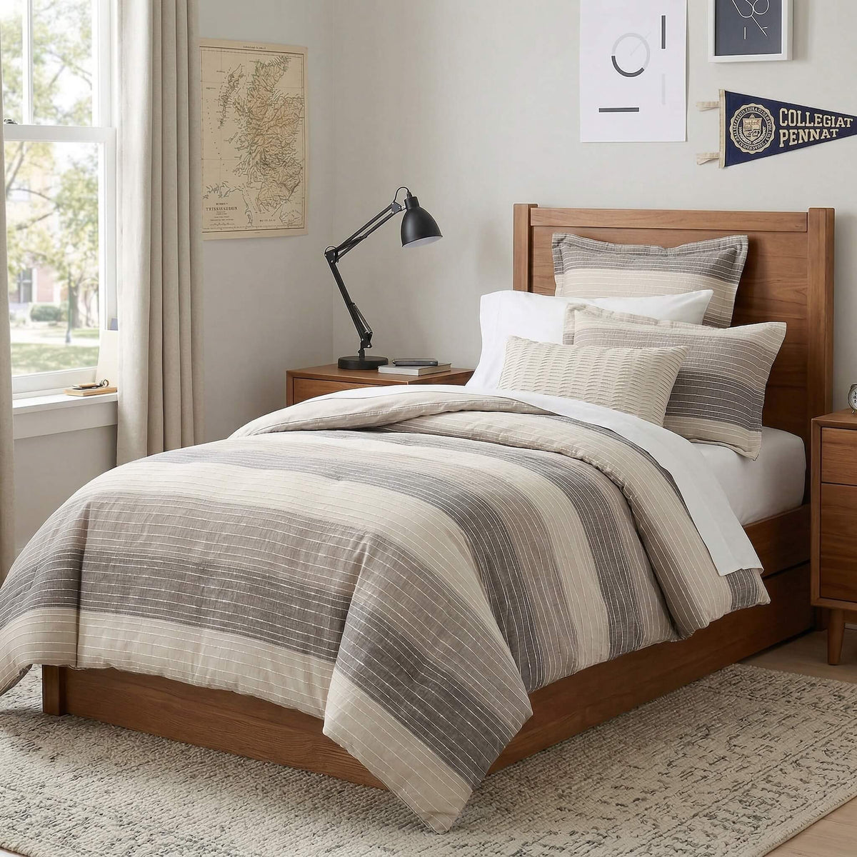 A wooden bed frame dressed with a striped beige, gray, and white Twin XL bedding set, styled in a bright, neutral-toned dorm room with a nightstand, desk lamp, and framed wall art.