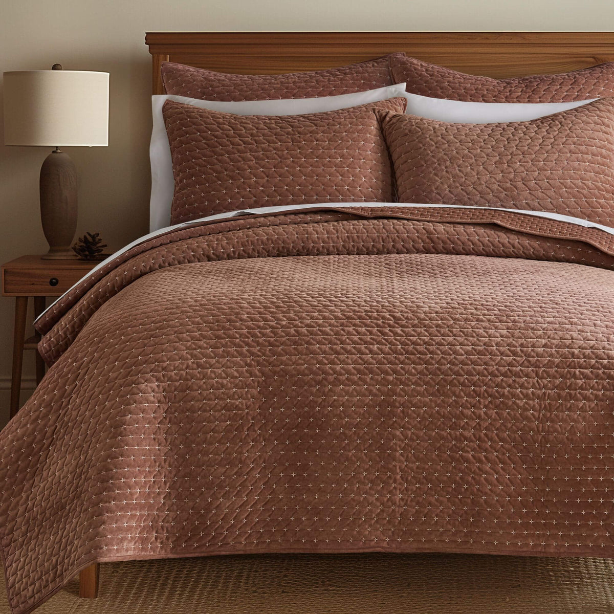 A bed dressed in a quilted dusty rose-brown bedding set with matching pillow shams, white pillows, a wooden headboard, and a bedside lamp.