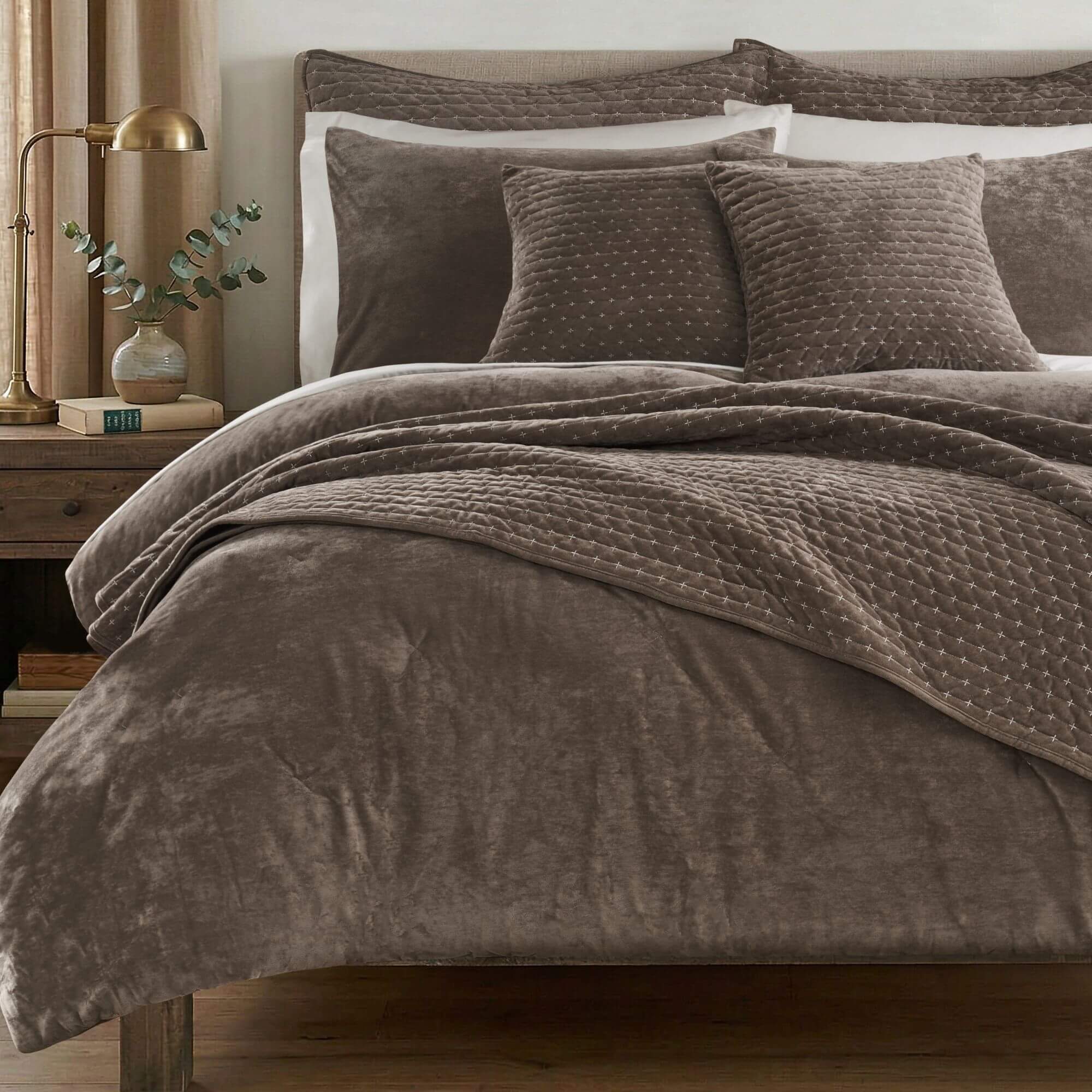 A bed dressed in a plush taupe-brown quilted bedding set, including a comforter, matching pillow shams, and two decorative throw pillows, styled in a warm bedroom setting with a wooden nightstand and brass lamp.