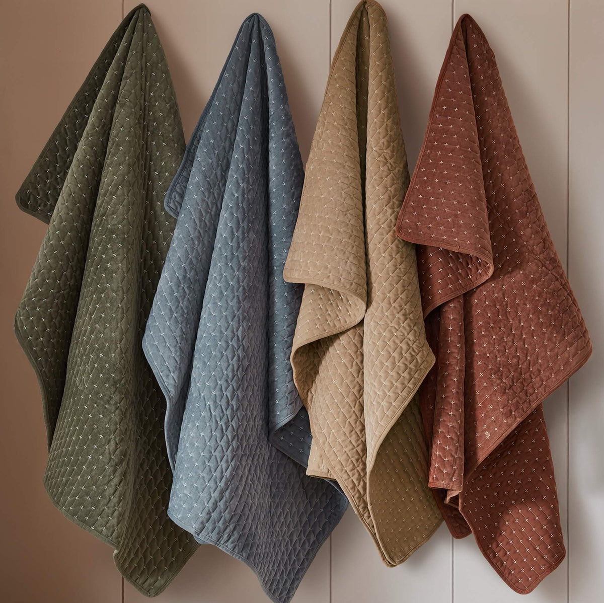 Four quilted velvet throws in muted earthy tones hanging on a light wood paneled wall. 