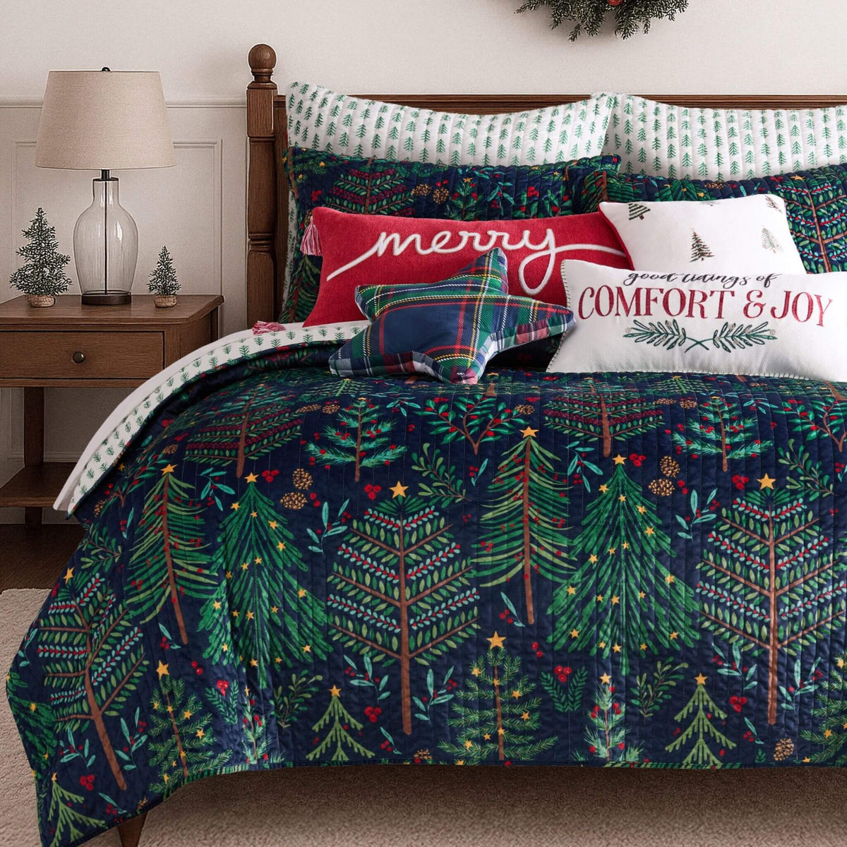A festive Christmas-themed bedding set on a bed, featuring a dark navy quilt with a green pine tree pattern, matching patterned pillows, and accent pillows with holiday text and plaid designs.